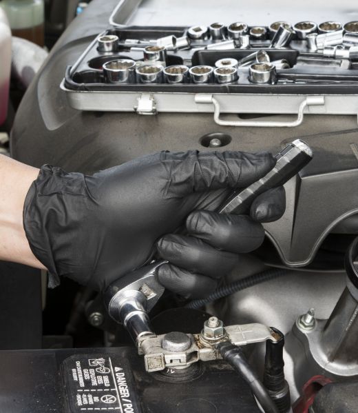 A mechanic tightens a battery terminal cable to a lead battery post after cleaning it with a wire brush during routine automobile maintenance.
